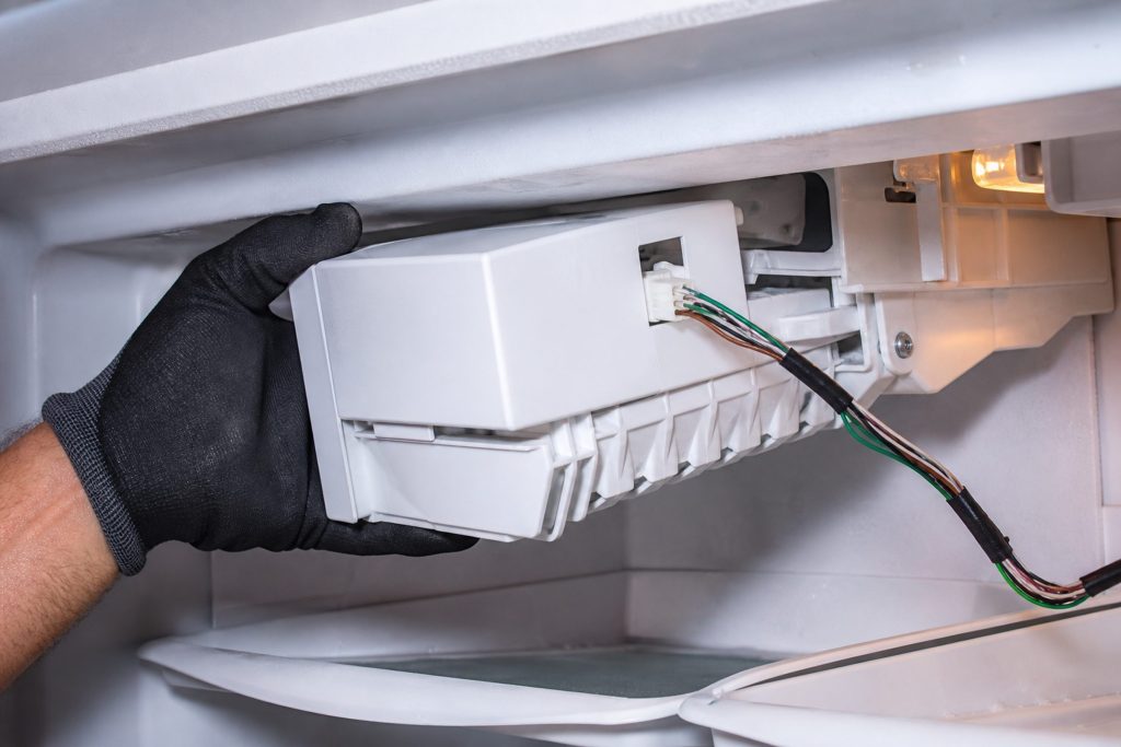 Gloved hands drawing an ice maker module out of a freezer compartment with its wiring loom still attached
