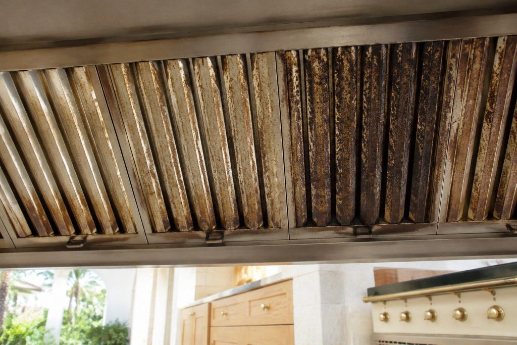 The angled baffle filters inside a hood canopy seen from directly below, their steel dulled and furred with a brown film of old grease, one filter darker than the rest.